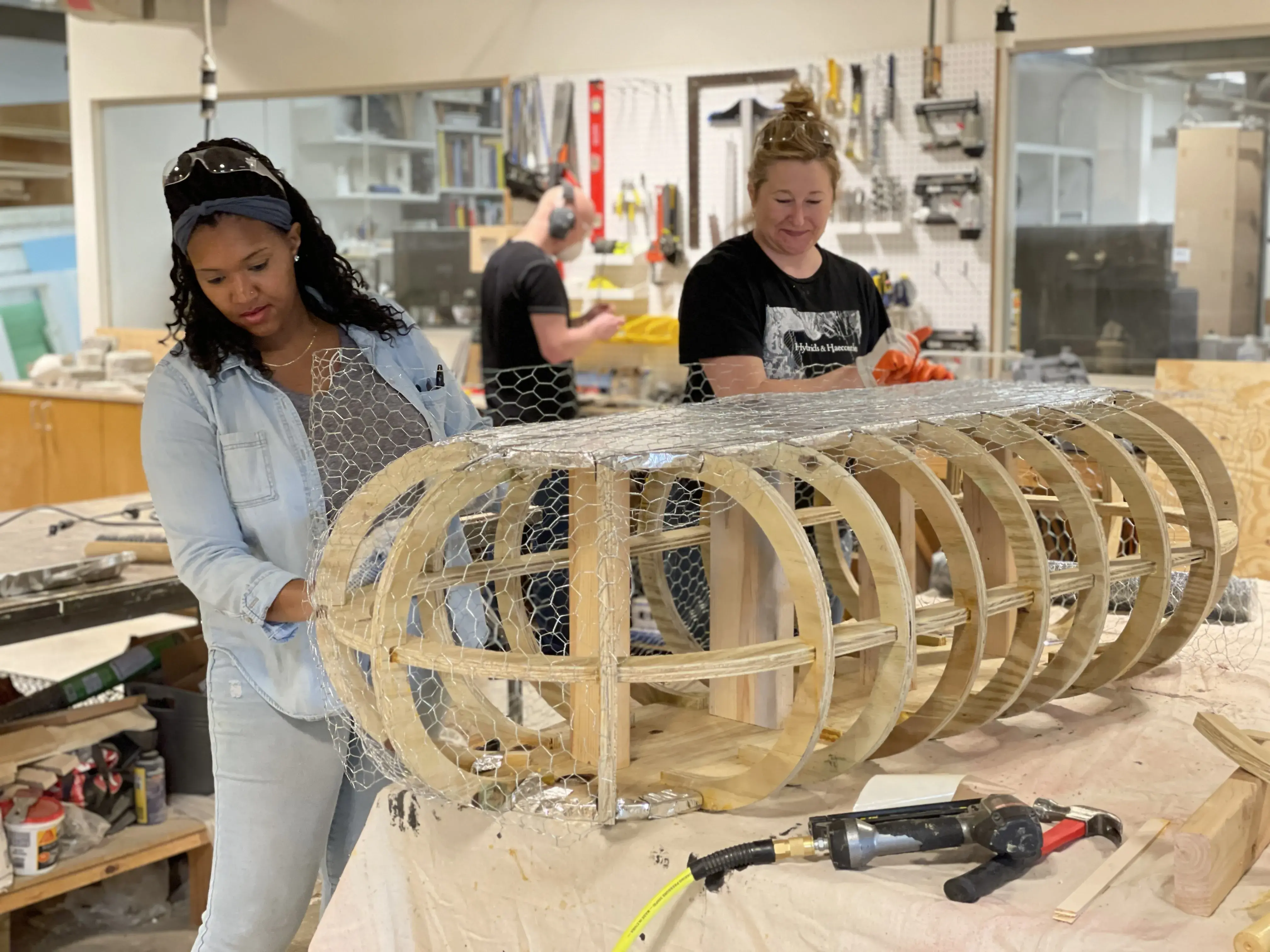Two women are fitting chicken wire around a rounded wood frame.