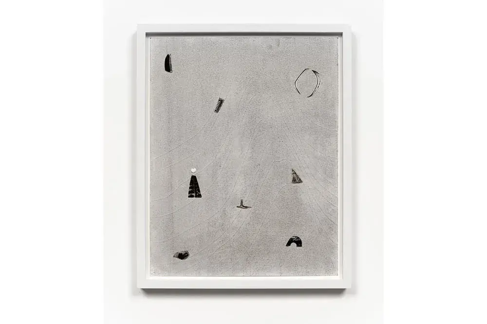 A grayscale artwork with small darker items on a lighter background hangs in a white frame