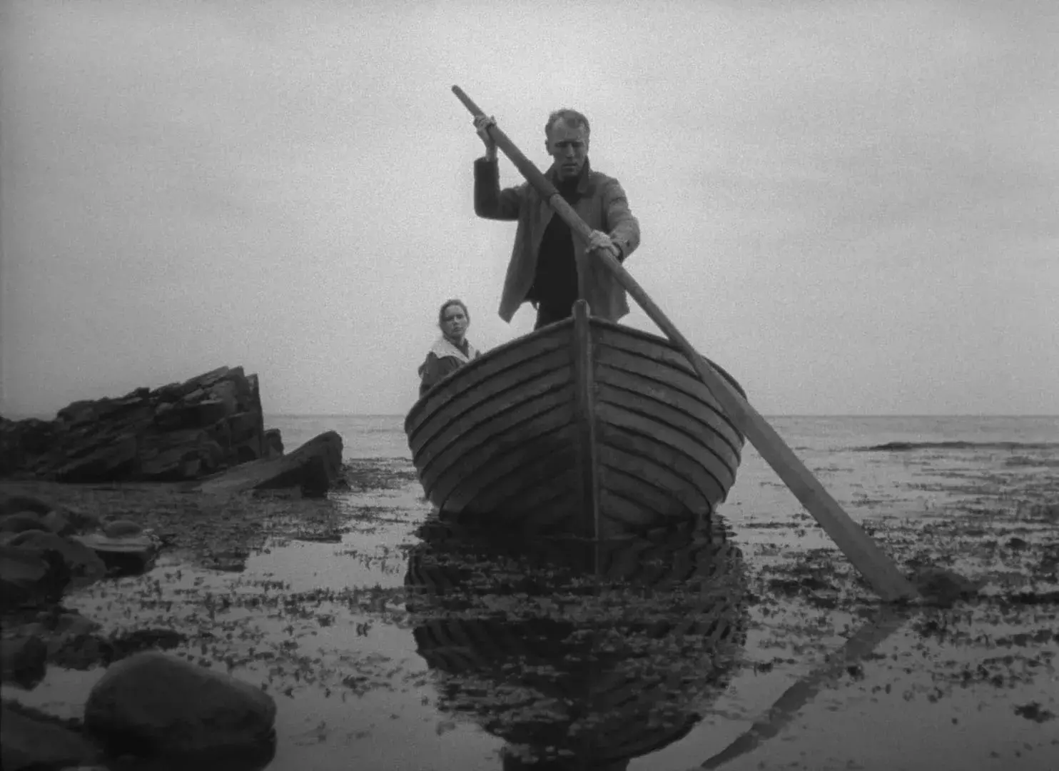 A black and white image of a man rowing a large boat to shore. A woman is sitting in the back of the boat.