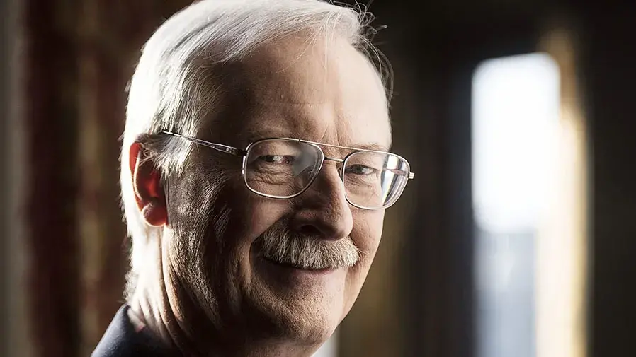 John Musker smiles at the camera. He has white hair and a gray mustache and wears glasses.