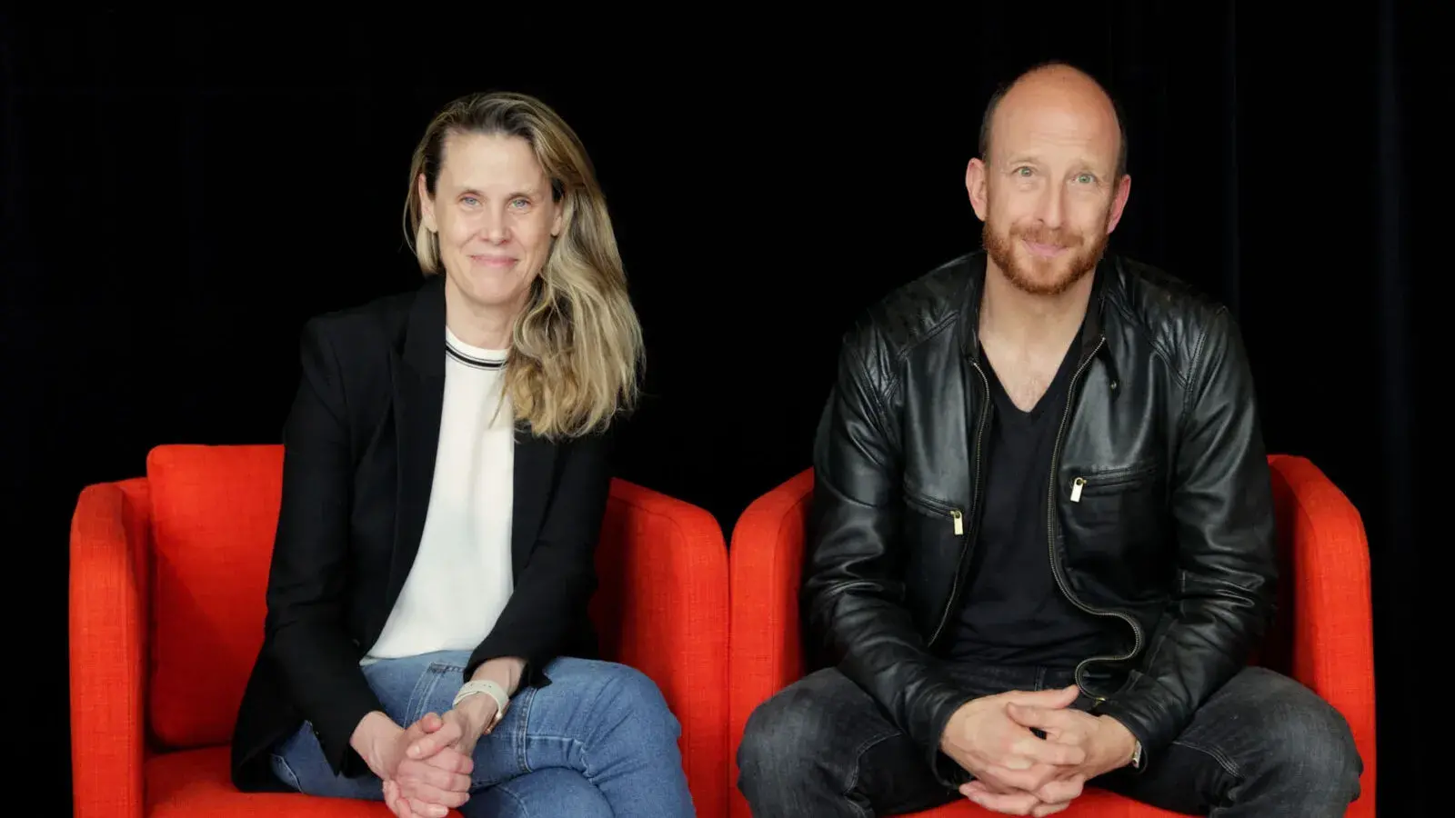 A blond woman and a bald man with facial hair sit on red chairs next to each other