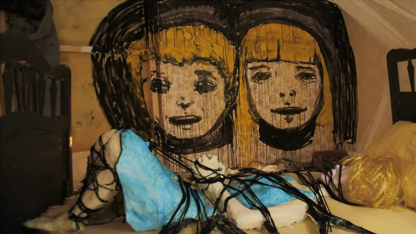 A puppet of a woman is tied to a bed. On the wall above her are the painted faces of two children. 