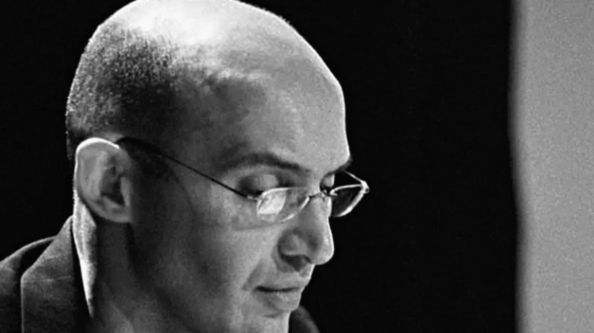 Walid Raad looks down at an angle from the camera. He has a bald head and wears glasses