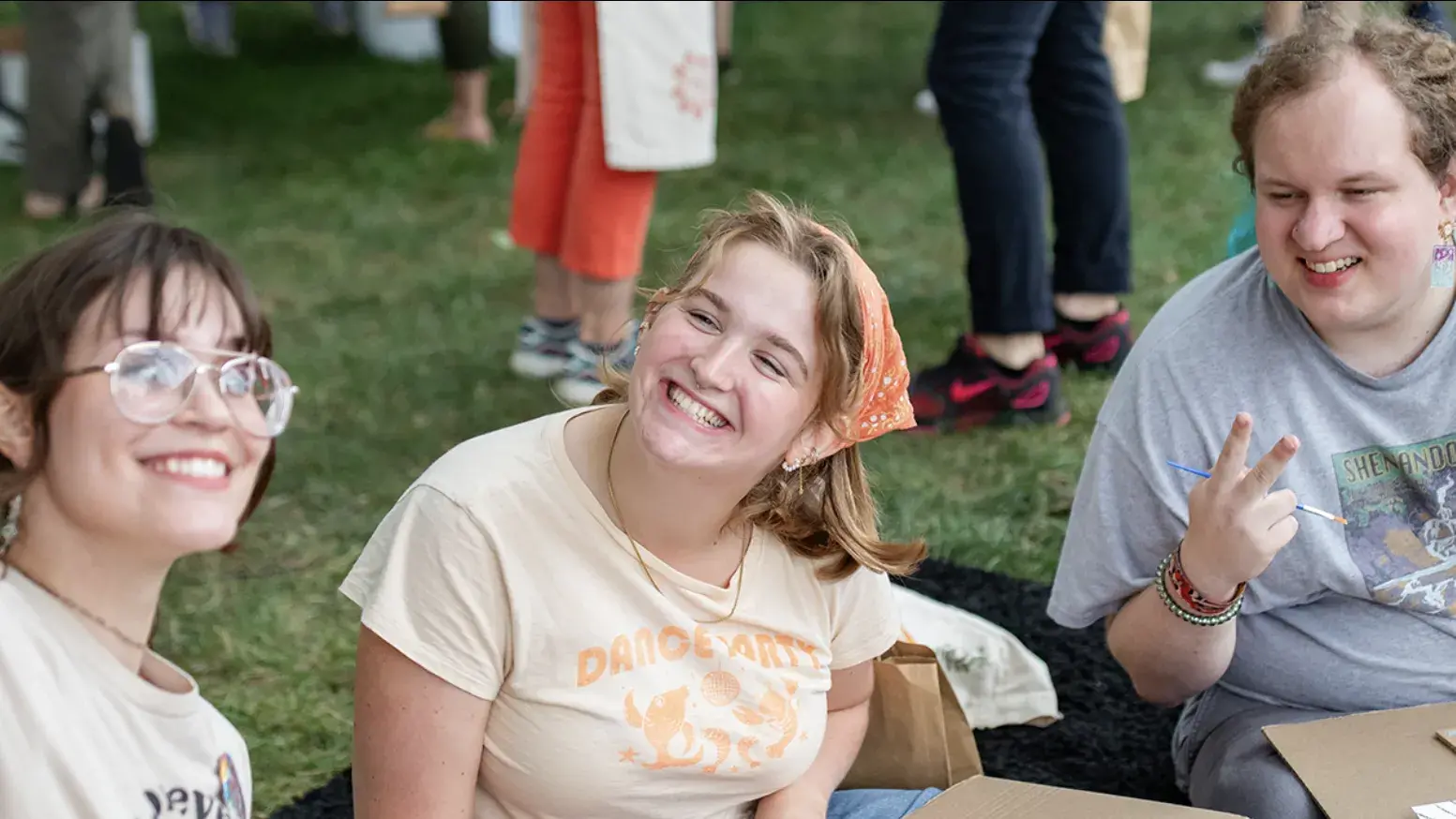 Three adults sitting down on the grass taking a selfie.  