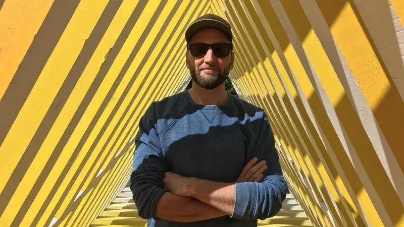 David Buckley Borden stands in a triangular space and smiles at the camera wearing a baseball hat and sunglasses