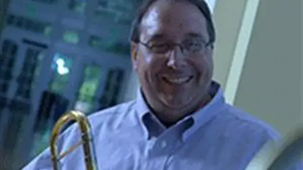 Bruce Tychinski smiles towards the camera and holds a trombone which is mostly not visible in the photo