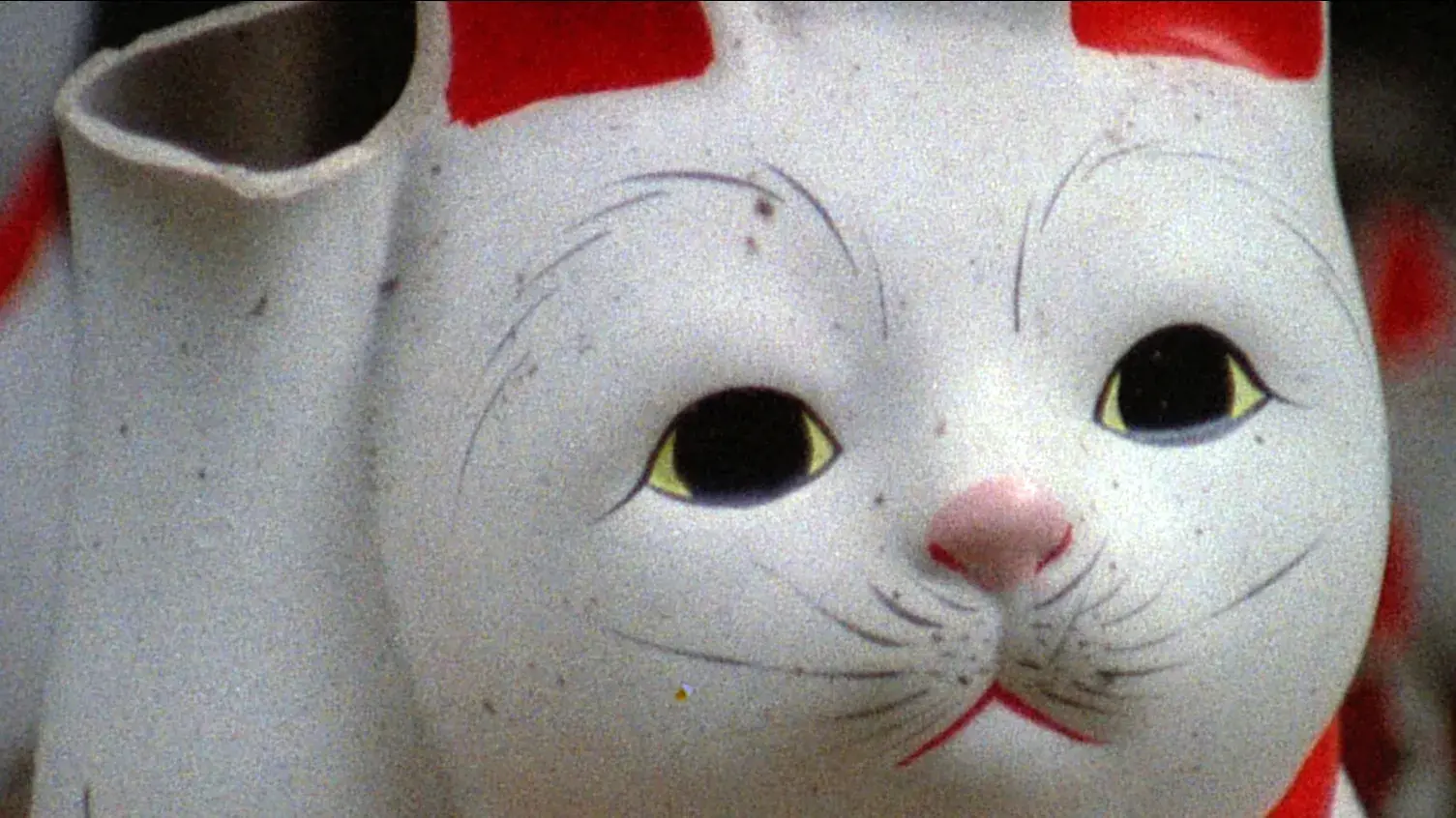 A closeup of a white cat sculpture with painted red ears, a red collar, and whiskers. The end of its raised paw is broken off.