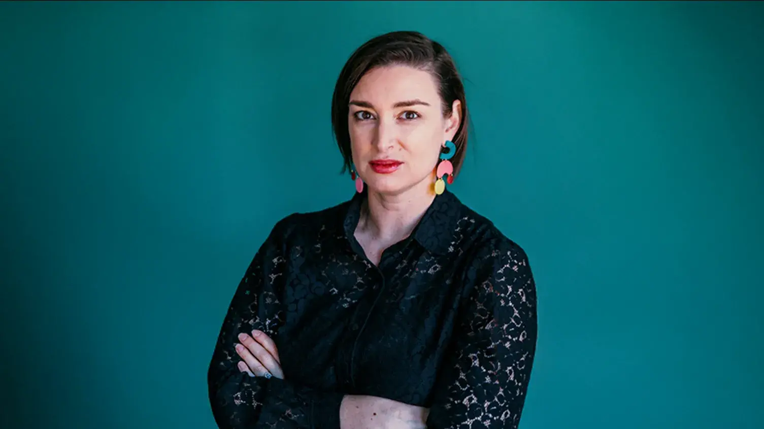 A headshot of Leah Triplett, who has light skin and short brown hair. She wears a black blouse and colorful earrings.
