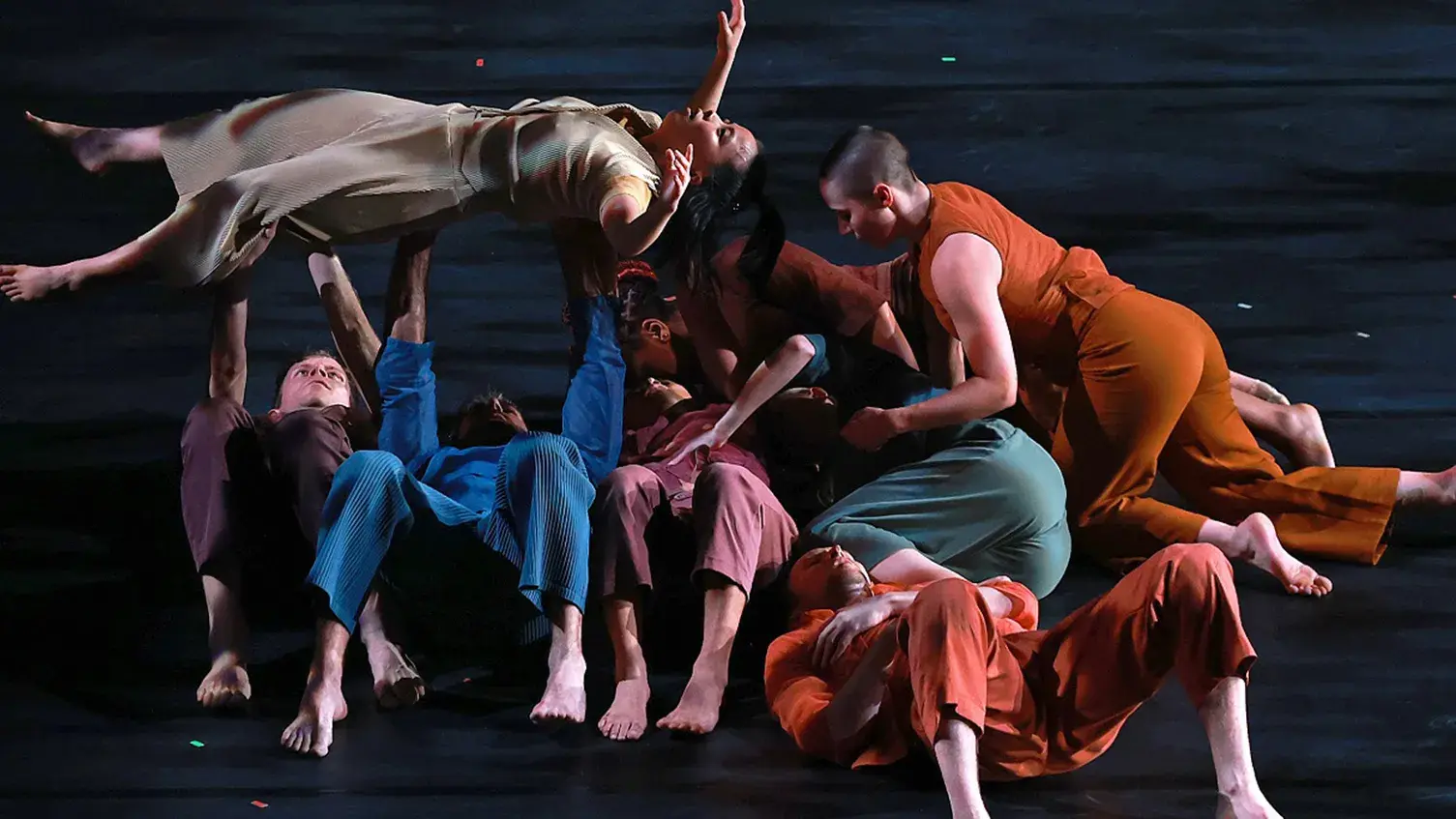 Multiple people lie on a stage and several of them are holding up a person who is lying down