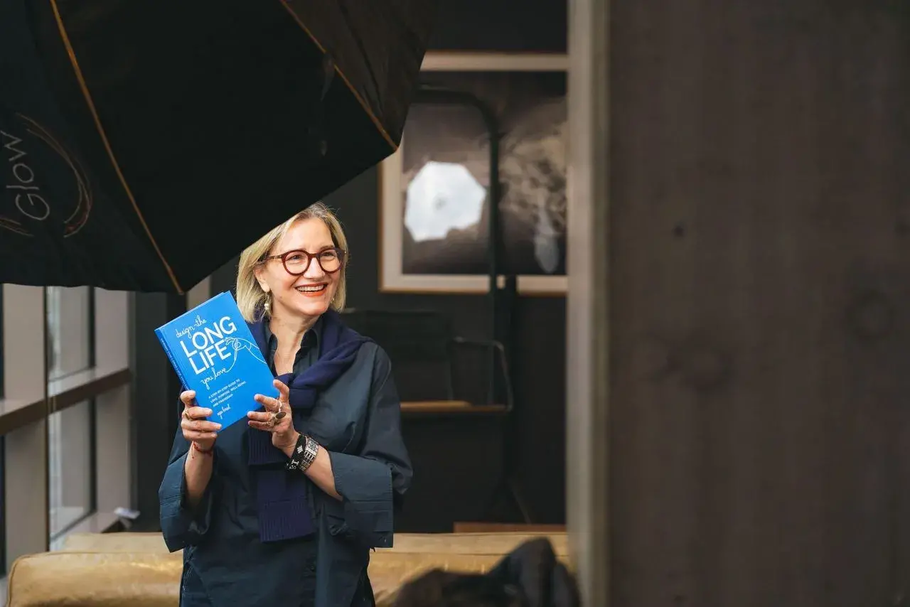 A woman wearing glasses with straight light-colored hair holds a bright blue book and smiles