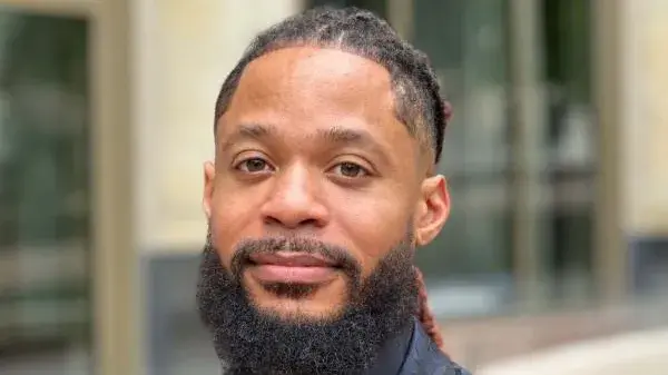 Photo of Terron Banner, a man with medium colored skin, dark hair pulled back and facial hair. He wears a dark blue and black suit jacket and shirt and looks at the camera.
