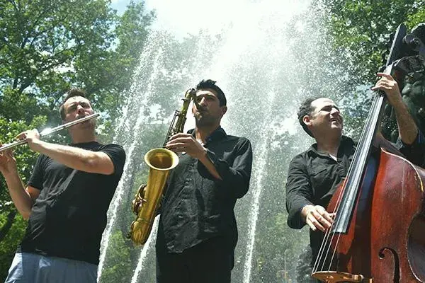 Three men playing instruments in front of trees. One man is playing the flute, one is playing the saxophone, and the final man is playing the double bass.