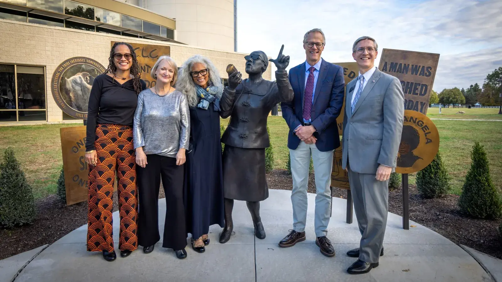 Several people stand amidst and outdoor bronze sculpture of a woman speaking