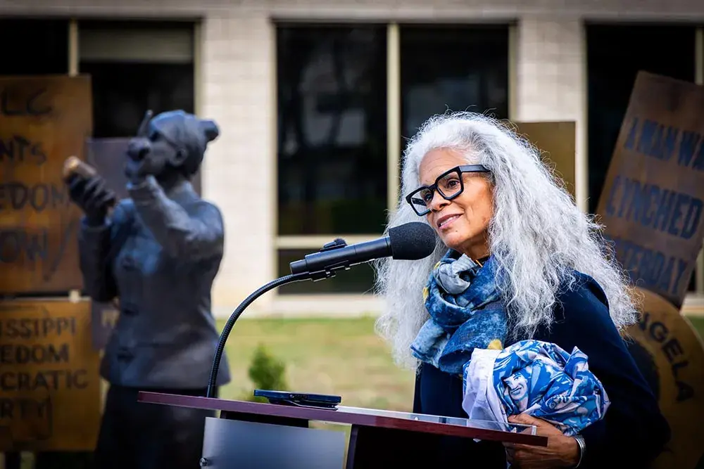 Artist Dana King speaks at a microphone in front of her statue of Ella Baker