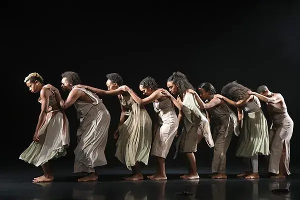 Multiple people stand in a line on stage with their hands on the shoulders of the person in front of them