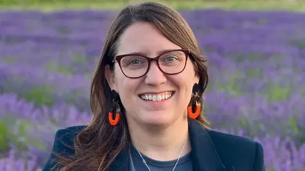 Headshot of Lauren Tilton, a woman wearing glasses and bright earrings standing in front of a field of purple flowers