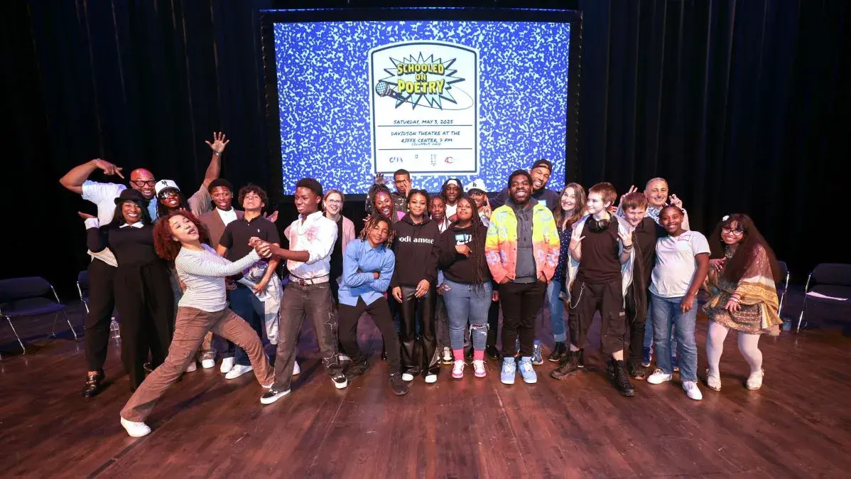 A group of students stands on a stage in front of a projected image of a composition notebook.