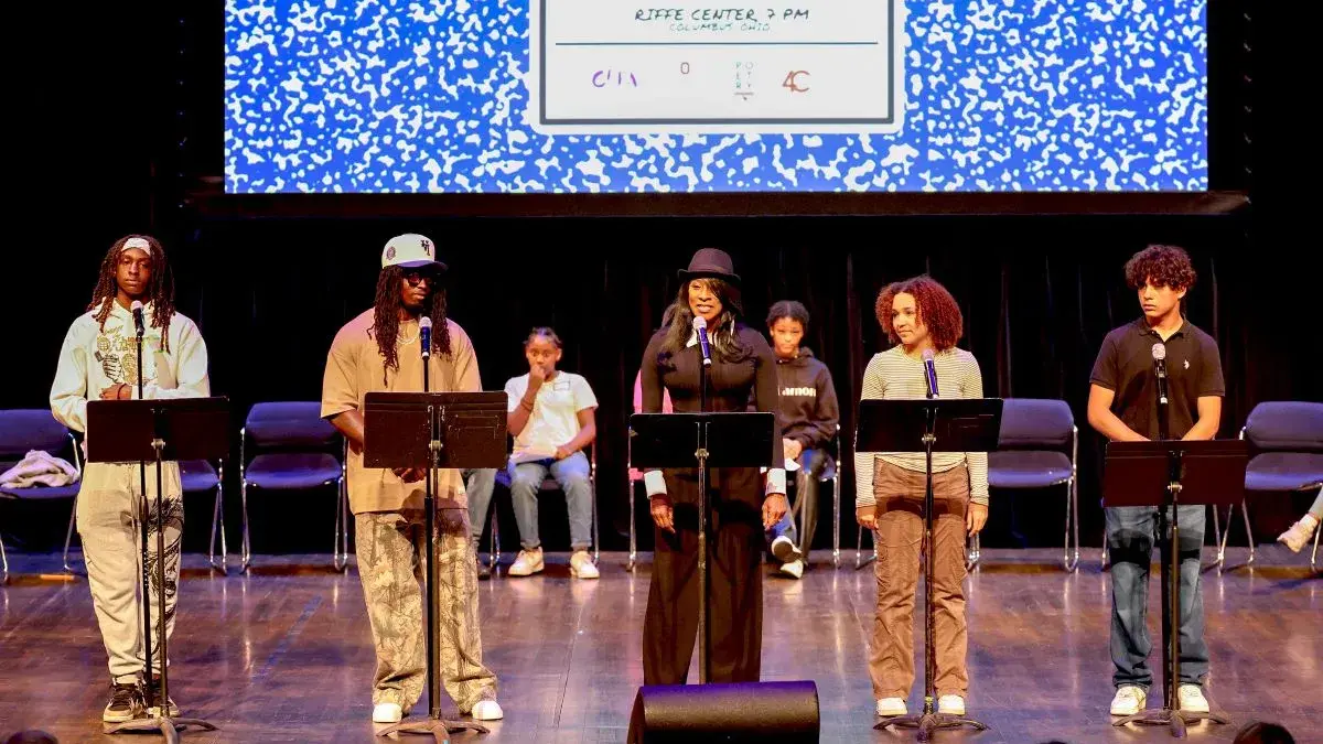 A group of students stands on a stage and read from materials on music stands. There is a projected image of a composition notebook on the screen behind them.