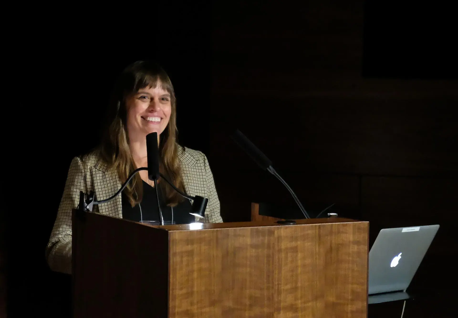 Kate Shannon, a white woman with long straight dark hair, stands behind a podium in a dark space