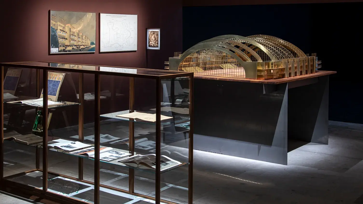 A gallery containing a glass and wood display case, an architectural model of an open structure with curved roof supports, and images of a building and building plans.
