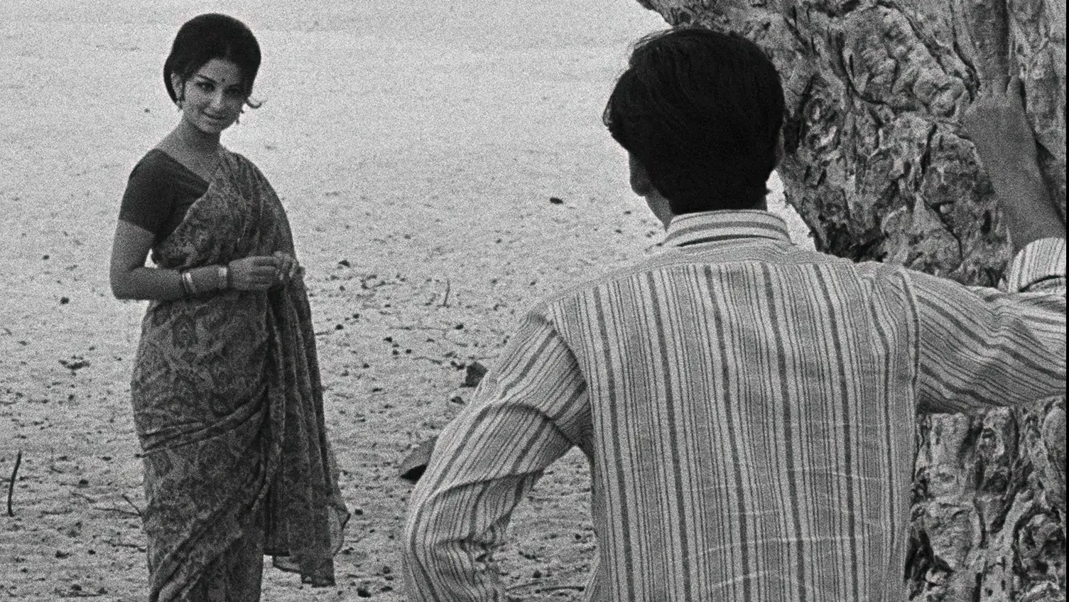 A woman wearing a sari stands in the sand, a man is facing her with his hand against a tree.