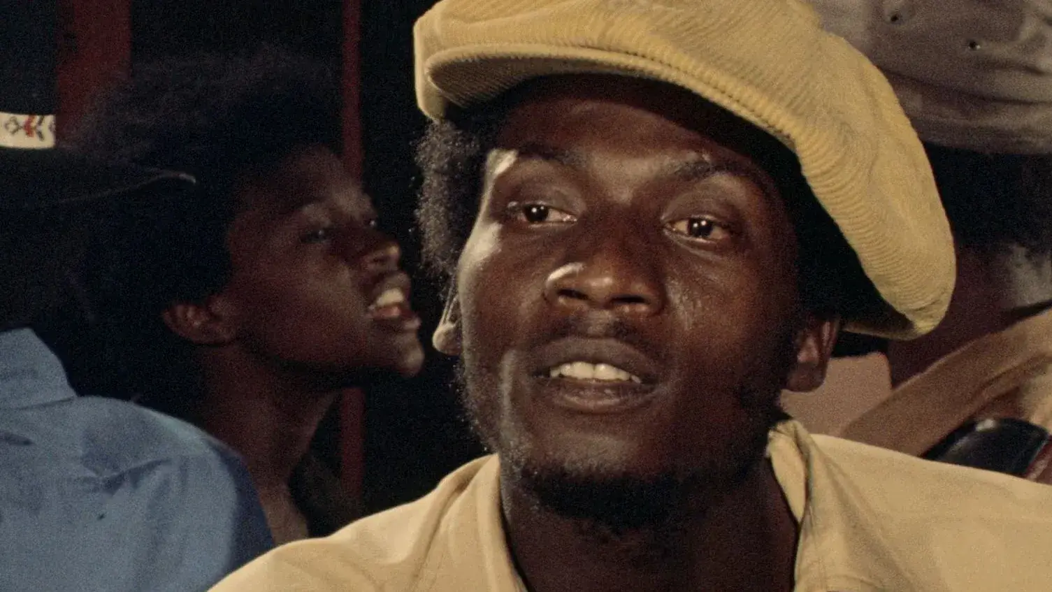 A man wearing a light yellow cap and shirt looks ahead, with other people visible in the background in a dimly lit setting.