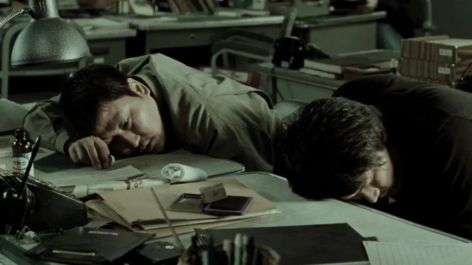 Two men lay their heads on a desk covered with papers in a dimly lit office.