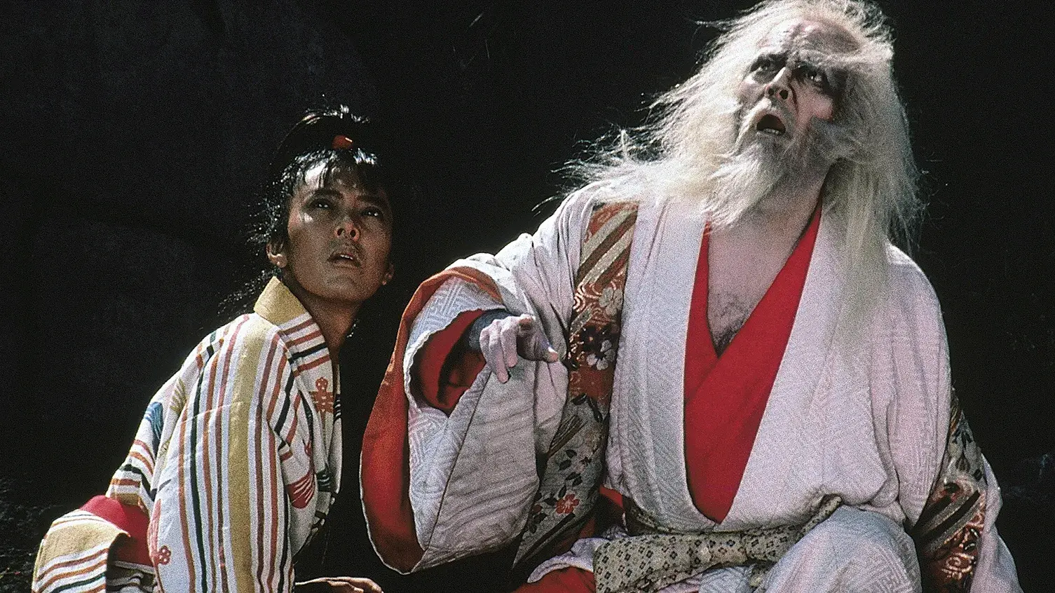 Two individuals in kimonos kneel on the ground together looking up at something.