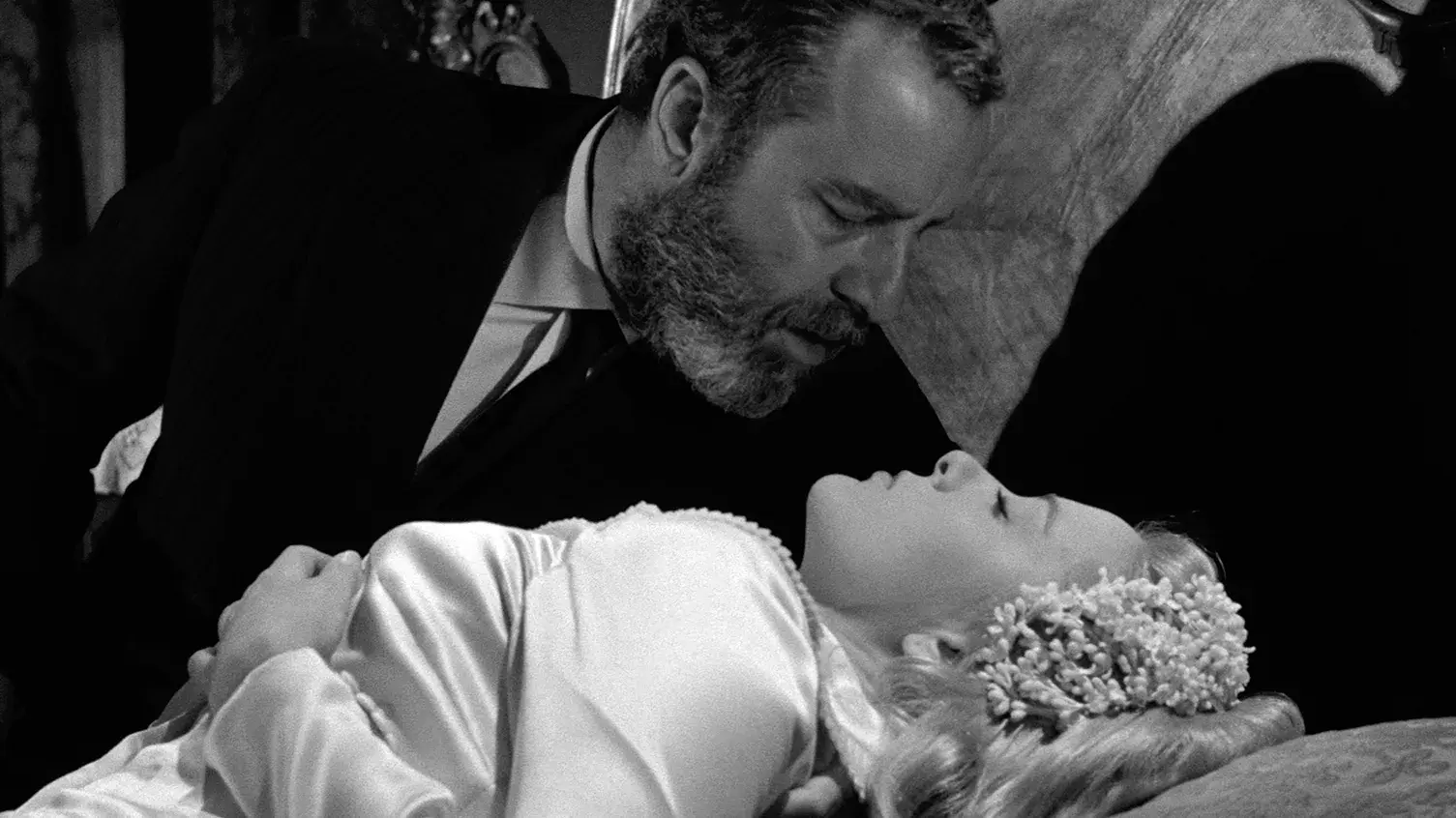 A bearded man leans over a woman in a wedding dress who is lying down on a bed, in a black and white scene.
