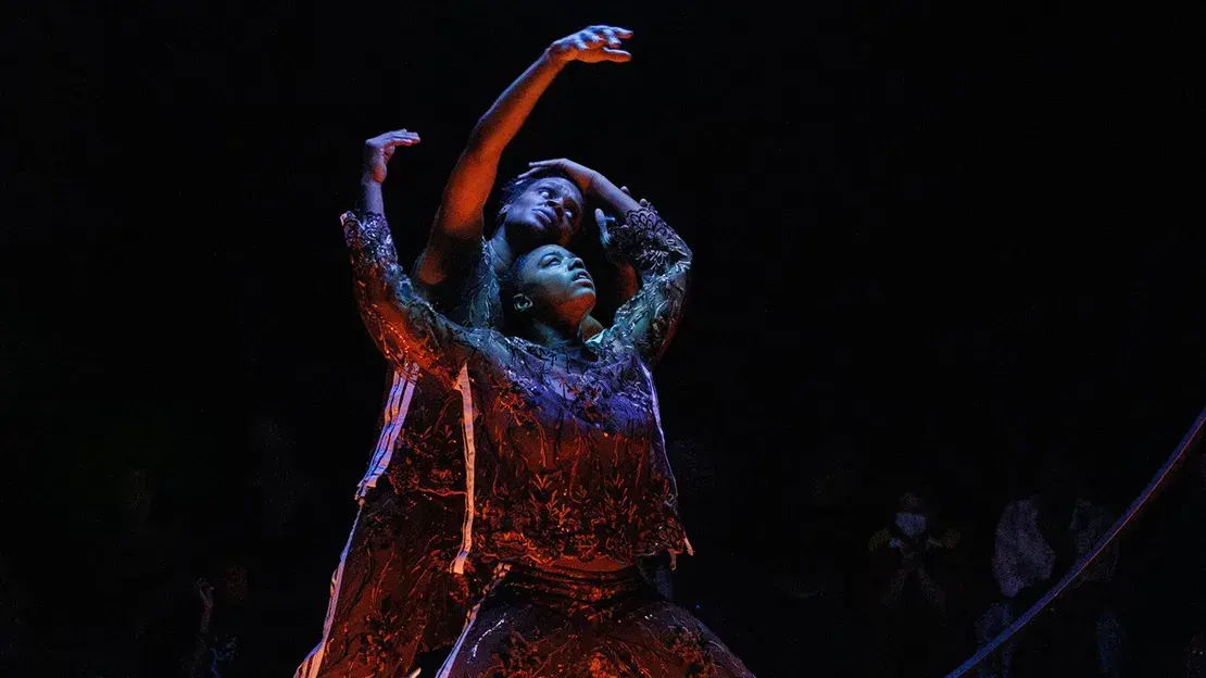 Two female dancers stand between two vertical metal rings on a dark stage illuminated from above by a blue disk.