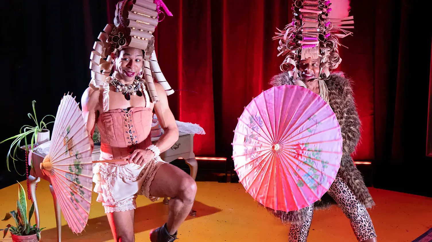 Two actors stand on stage wearing colonial wigs made of cardboard and holding parasols.