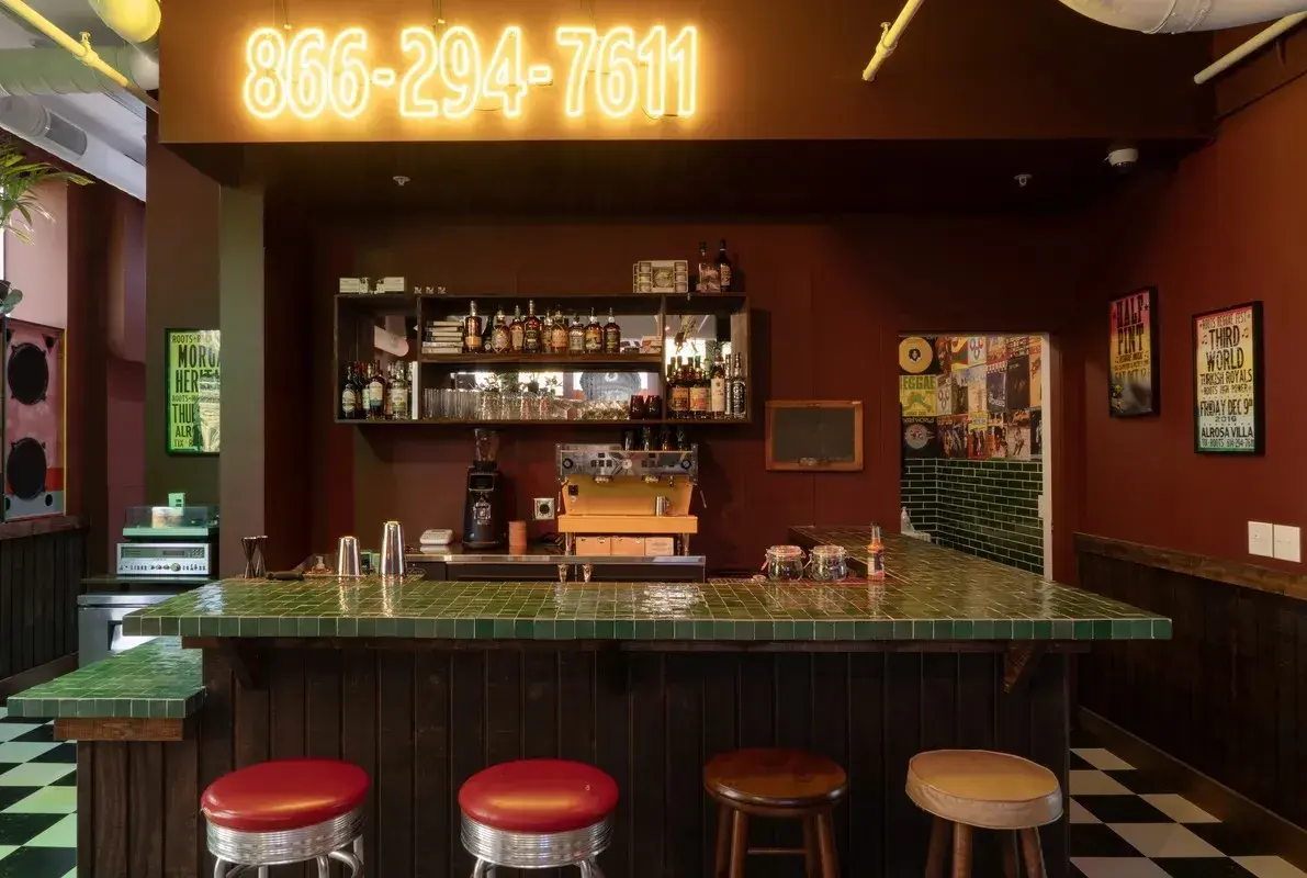Image of a small bar with a phone number above in neon and mismatched bar stools in front.