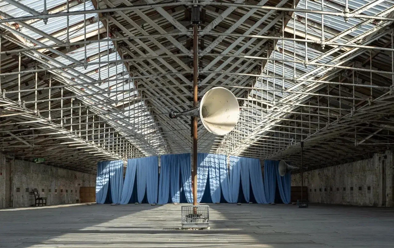 Blue curtains hang a the end of a large open interior hall. A megaphone is mounted on a pole in the foreground.
