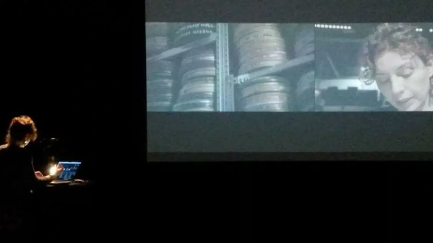 A person stands at a podium with a laptop in a dark room, watching a large screen displaying two film scenes: film reels and a woman's face.