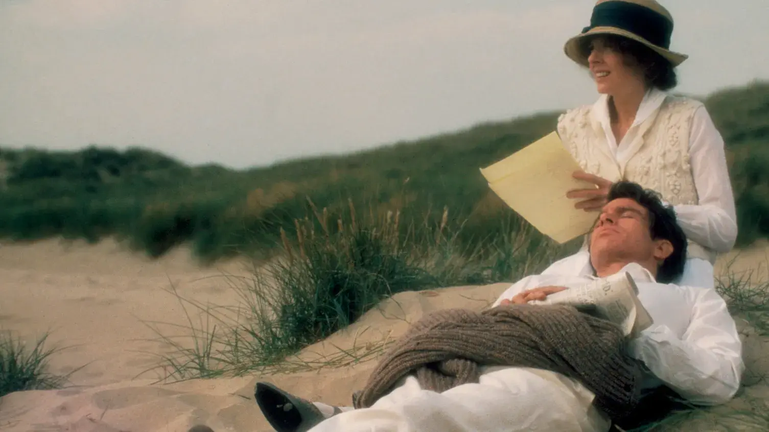 A woman sits while a man lies with his head in her lap. They both wear white clothes and hats, surrounded by grass on a sandy beach.