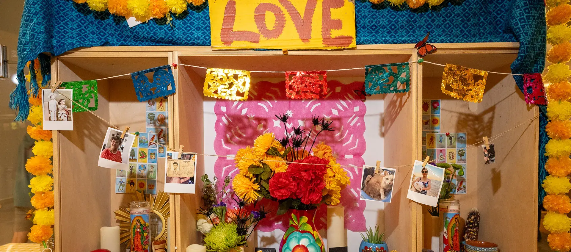Altar with paper and flower garlands, photos, candles, calaveras, vases, flowers, postcards, and a sign reading "Love".