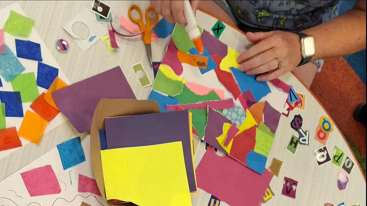 Overhead view of a tabletop featuring collages of colorful paper squares and of irregular torn paper shapes being glued down. 