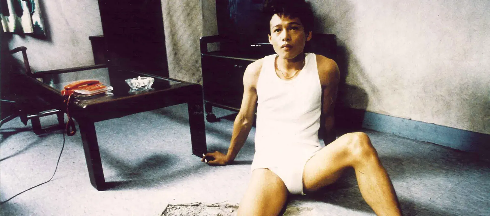 A man sits on the floor in his underwear and undershirt with his leg in a hole in the floor.