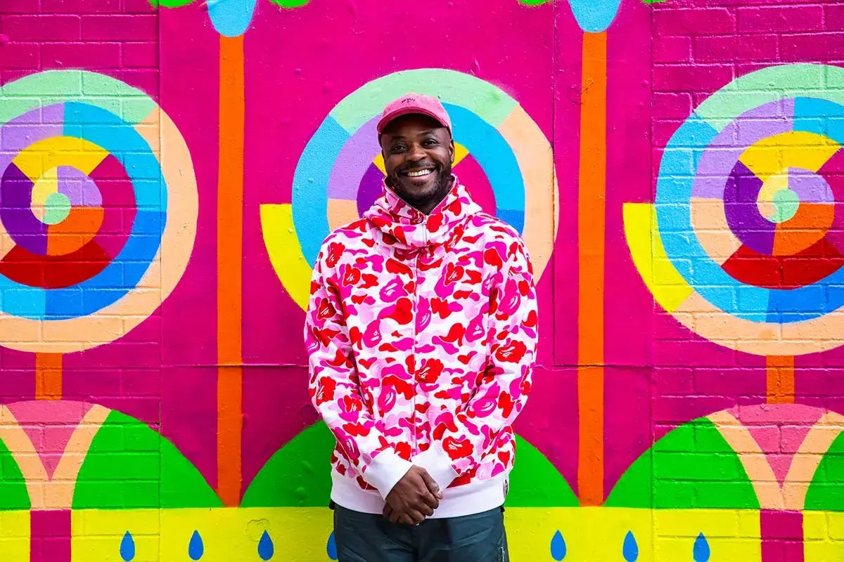 A smiling dark-skinned man wearing a hat stands in front of a brightly colored painted wall