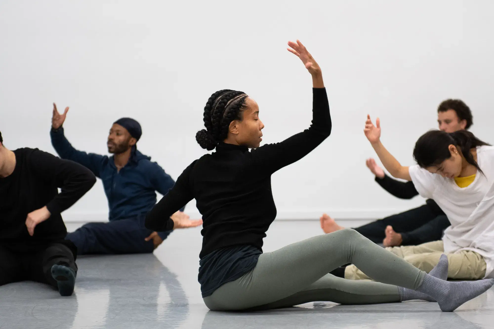 A Black woman wearing a black shirt and light green leggings sits on the floor and is in movement. Several other dancers are in movement near her.