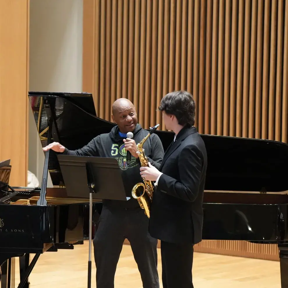 Brandford Marsalis speaks with a student holding a saxophone in front of a piano.