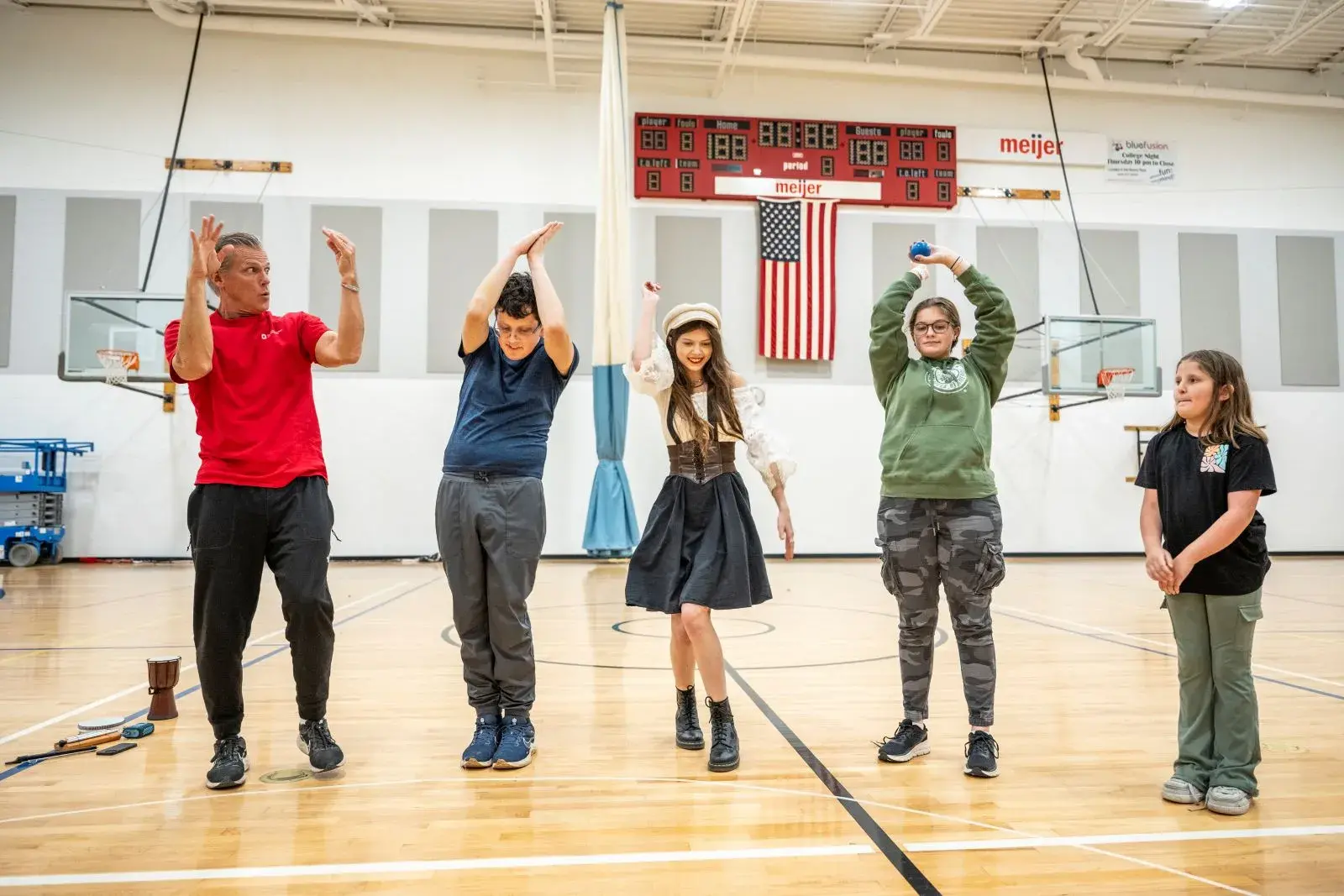 An adult and several young people stand on the floor of a gym and are moving their bodies in various ways