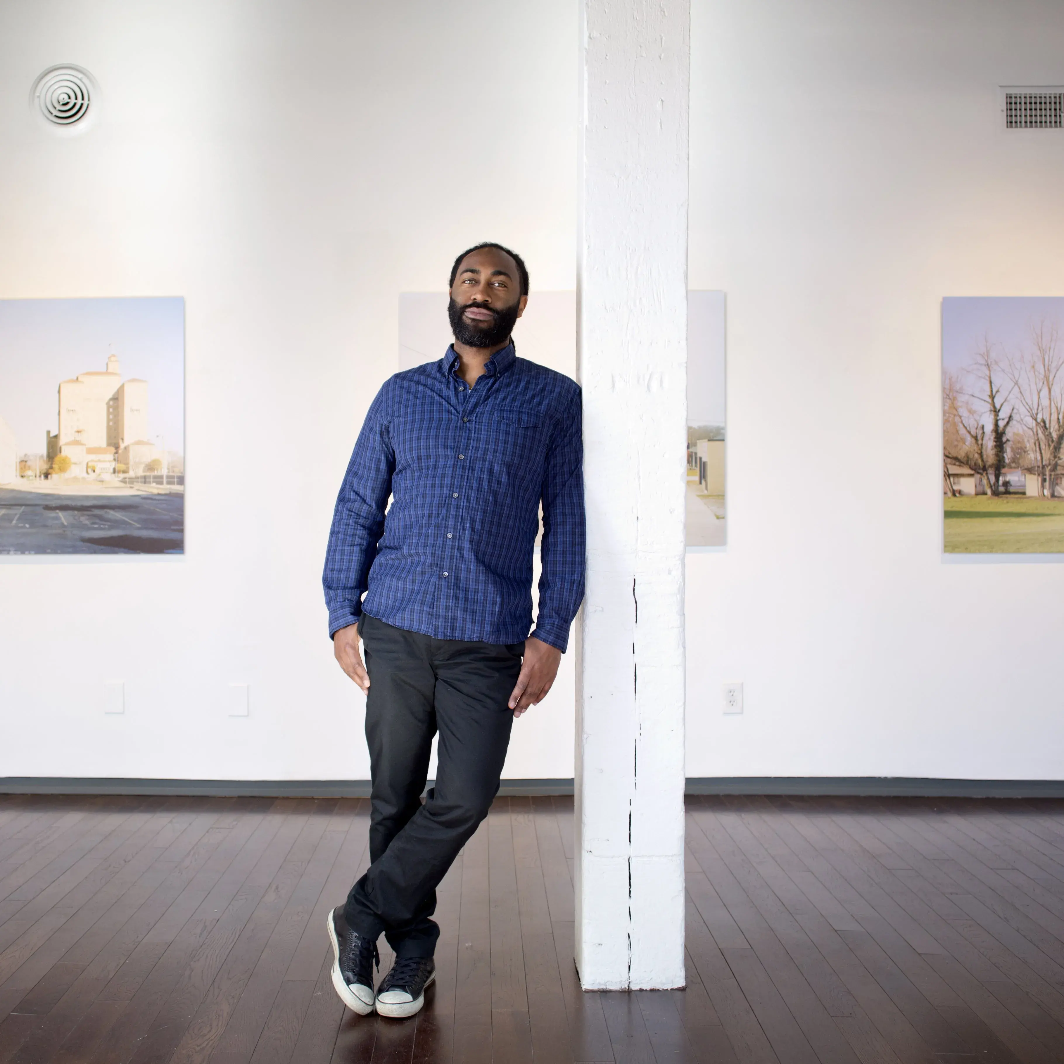 A Black man with a trimmed beard leans against a white pole. Several large photographs of buildings are hung on the wall behind him.