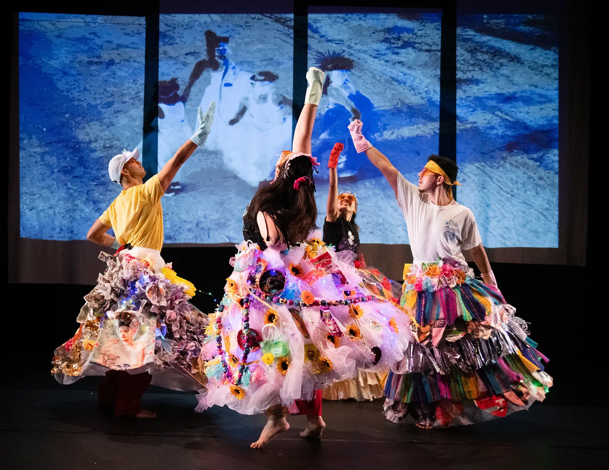 Four people wearing big multicolored skirts reach into the air in front of a blue projected background
