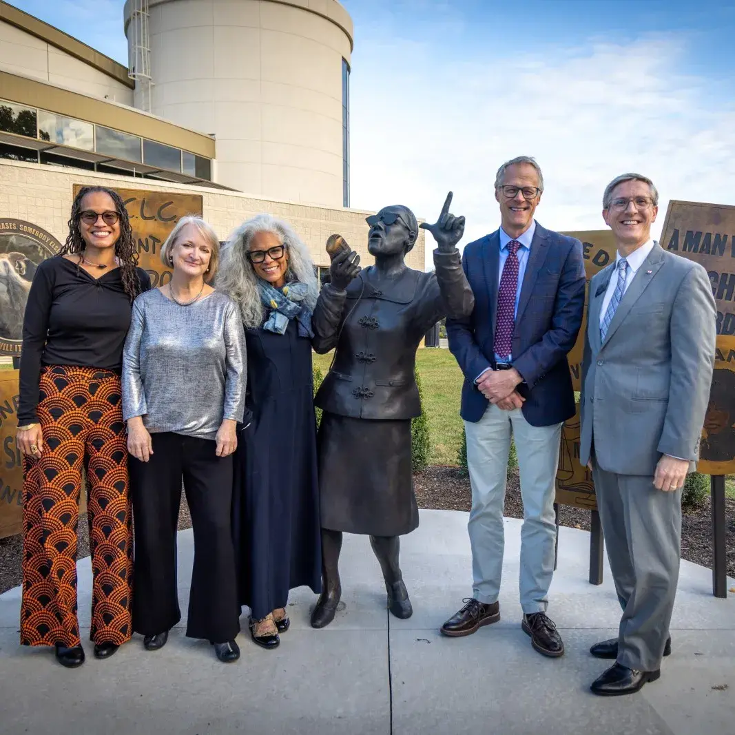 Several people stand amidst and outdoor bronze sculpture of a woman speaking