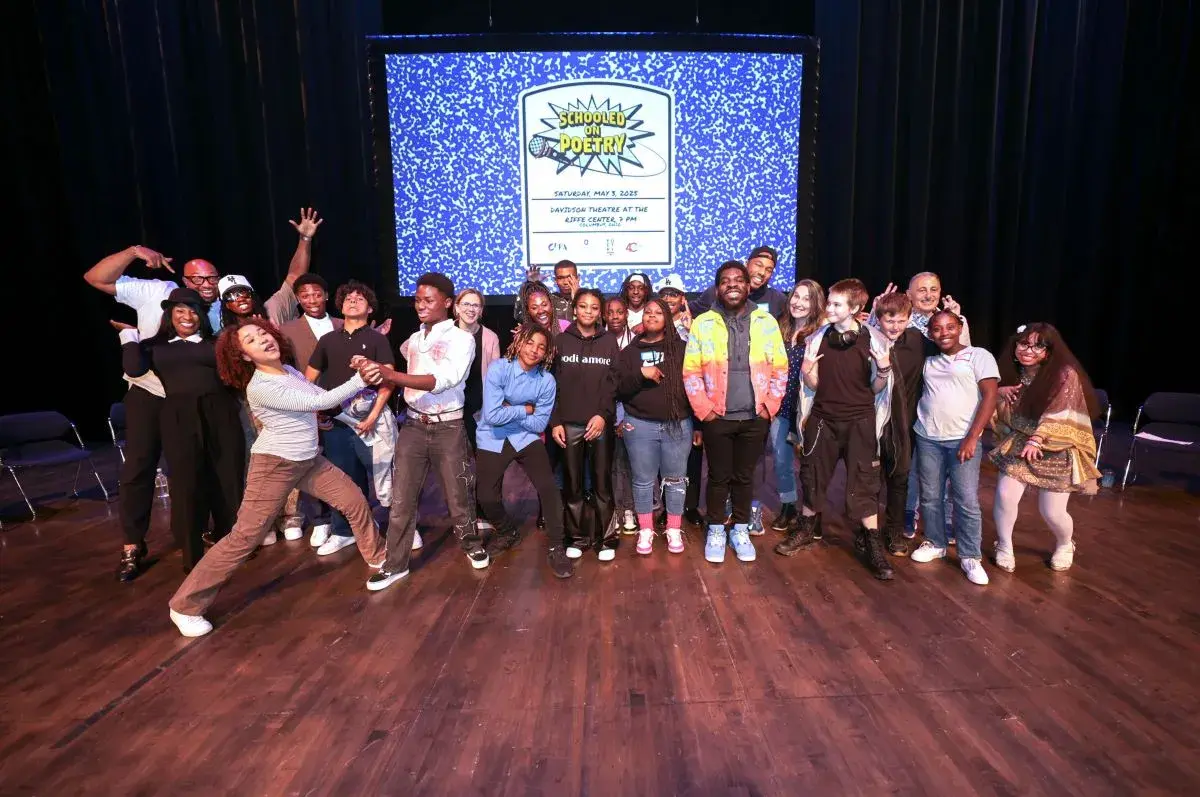 A group of students stands on a stage in front of a projected image of a composition notebook.