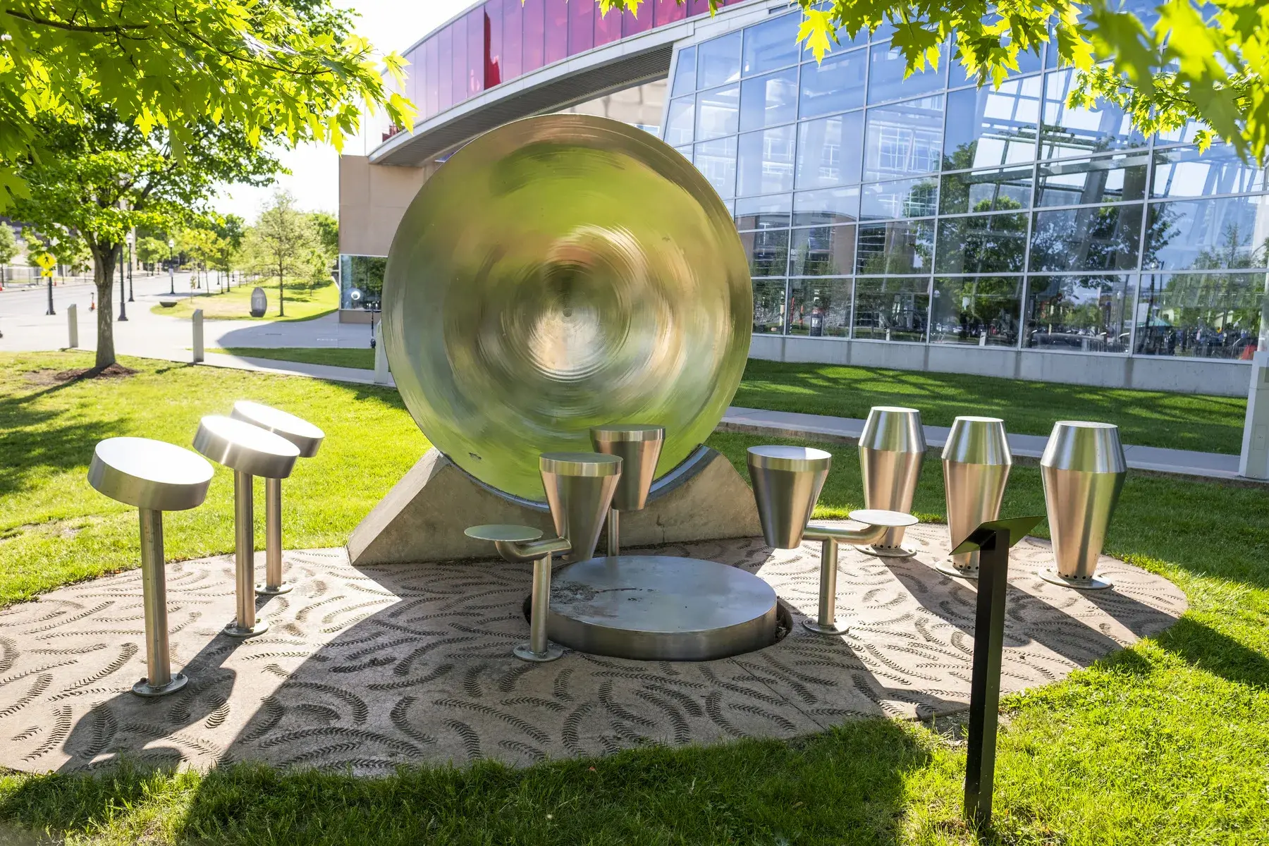 Multiple silver metal drums of various types sit on a textured area in the grass. There is a glass wall of a buildings behind it.