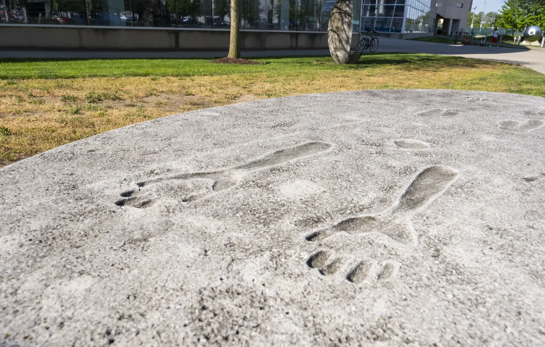 Top of a concrete object with an impression of a person's hands and forearms in it, with other impressions further back. An oval concrete bench is visible in the background.