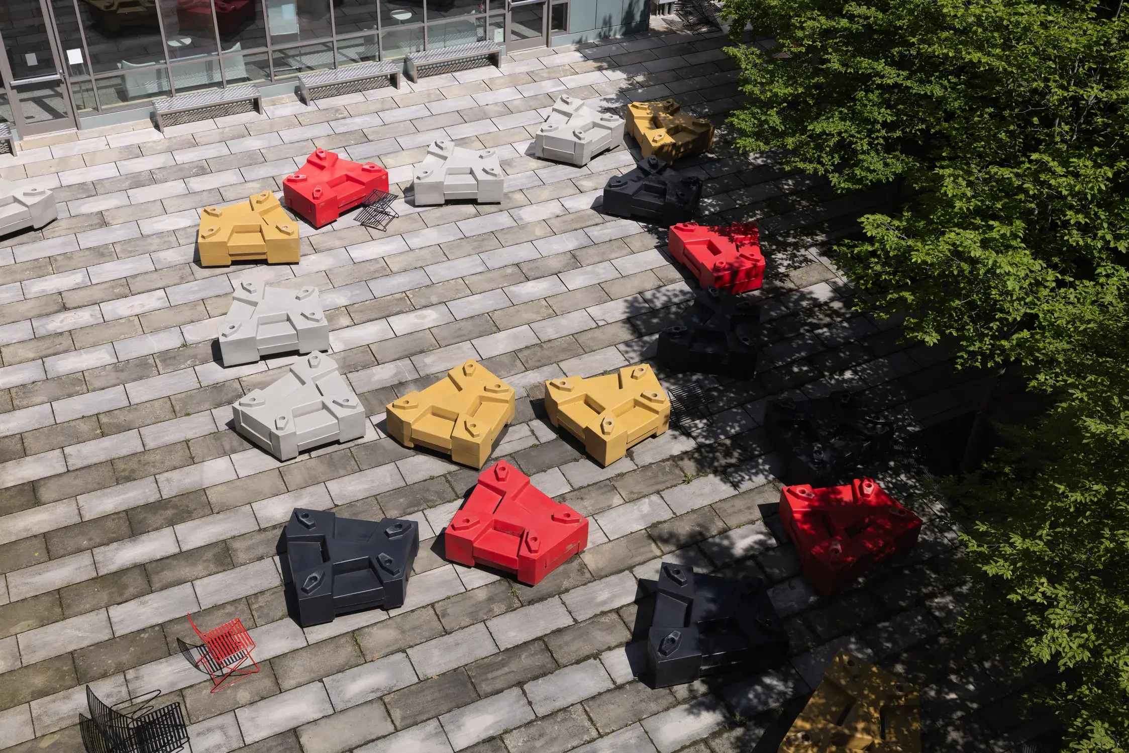 Red, yellow gold, gray, and black five-sided objects sit on a patio of light and dark gray pavers in alternating lines. There are trees on one side.