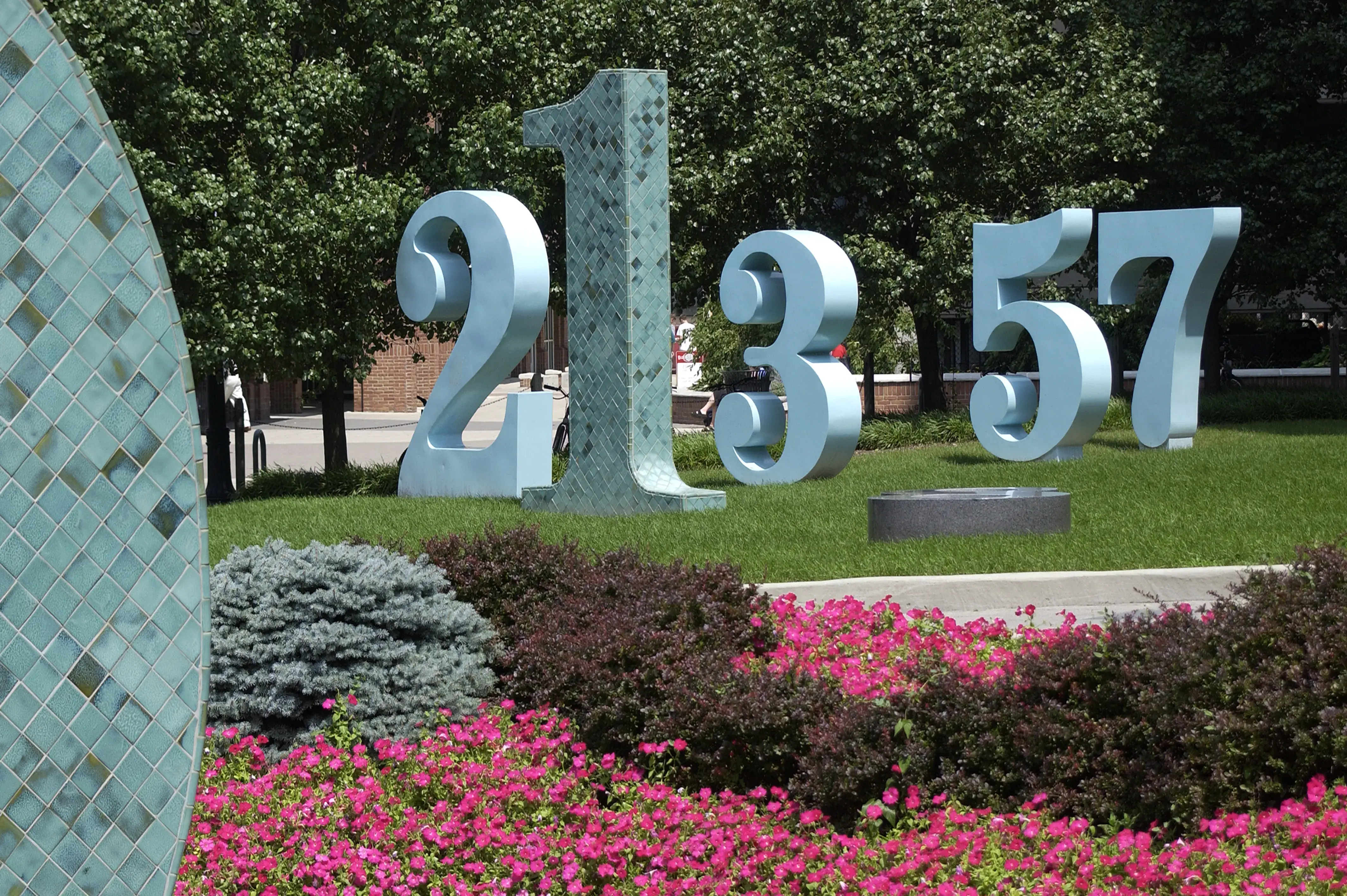 A group of large sculptural numbers in blue and tiles of green sit in a grassy area with flowers and bushes in the foreground. One number appears to be lying down in the grass.